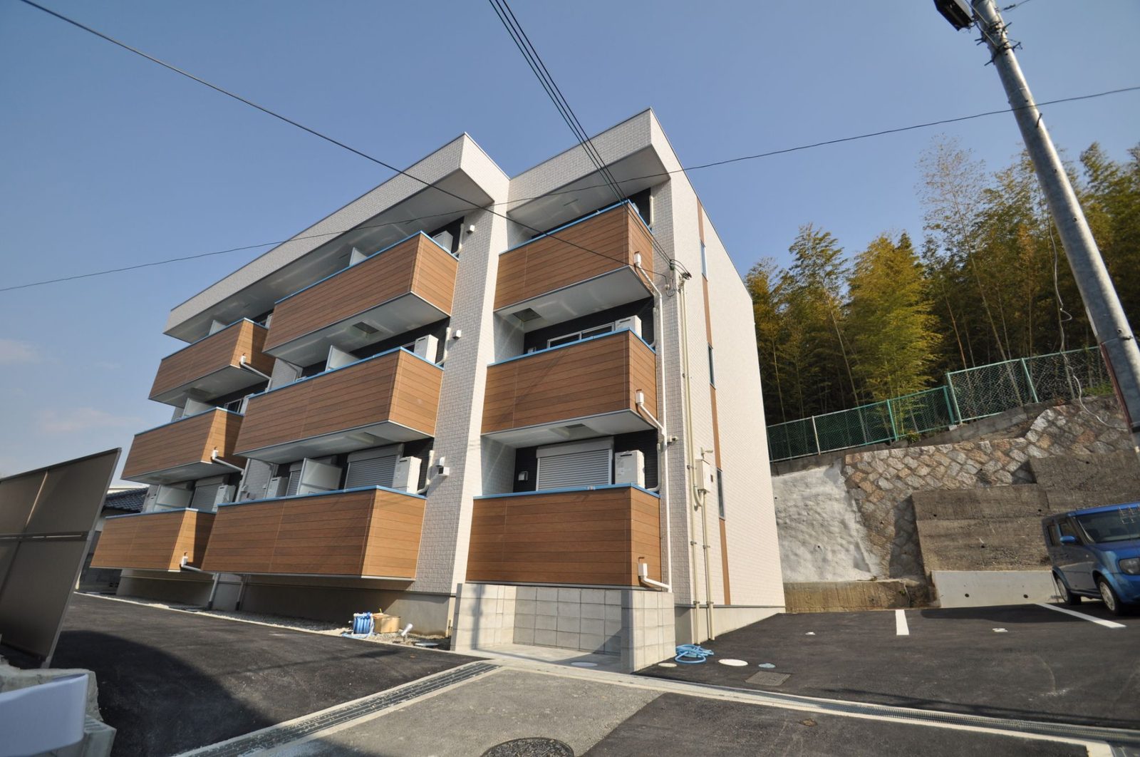 木目調のバルコニーが並ぶ白い3階建てアパート。背後に竹林と石積みの擁壁
